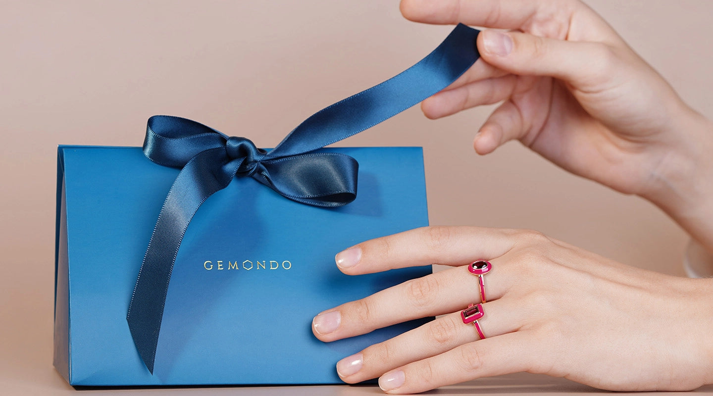 Blue gift box with a bow and 'GEMONDO' branding, held by a hand wearing a red ring.